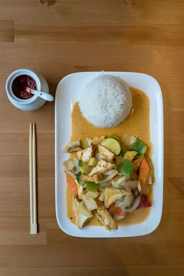 Red thai-curry with vegan tofu bits, zucchini, pepper, carrots and zucchini on a plate with rice, chop-sticks and a jar with chili paste on a wooden table - top view