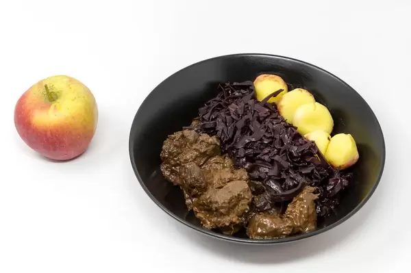 Rindergulasch nach traditioneller Art mit Apfelrotkohl und Miniknödel mit einem Apfel auf weißem Hintergrund