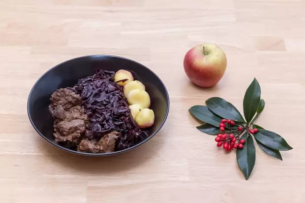 Rindergulasch nach traditioneller Art mit Apfelrotkohl und Miniknödel mit einem Zweig un einem Apfel auf einem Holztisch