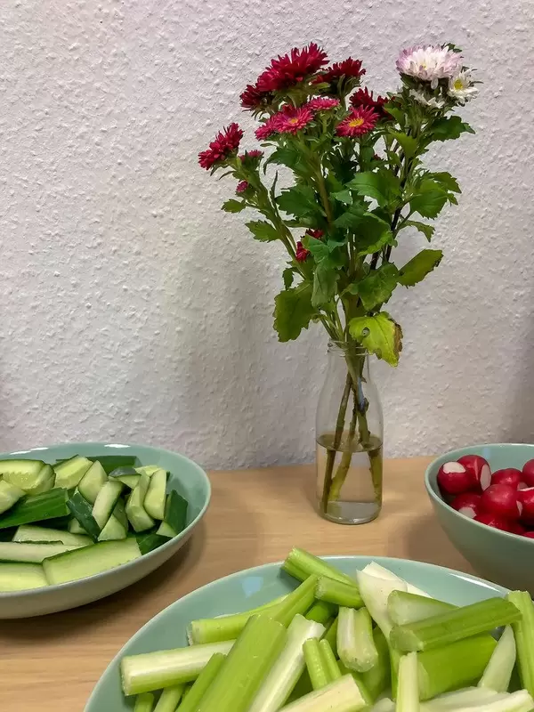 Rohkost - Gurken, Selleriestangen und Radischen in Schälchen mit einer Vase mit Blumen
