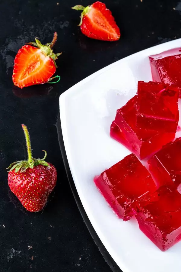 Rote Götterspeise-Stückchen auf einem weißen Teller, neben frischen Erdbeeren auf schwarzen Untergrund