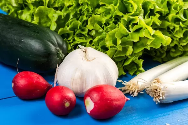 Rote Radieschen, Knoblauch, Gurke und Lauchzwiebeln vor Salatblättern auf blauem Holztisch