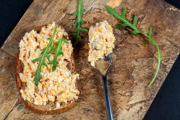 Rührei und Rucola auf einer Scheibe Schwarzbrot und eine Gabel mit Rührei auf einem Holzbrett