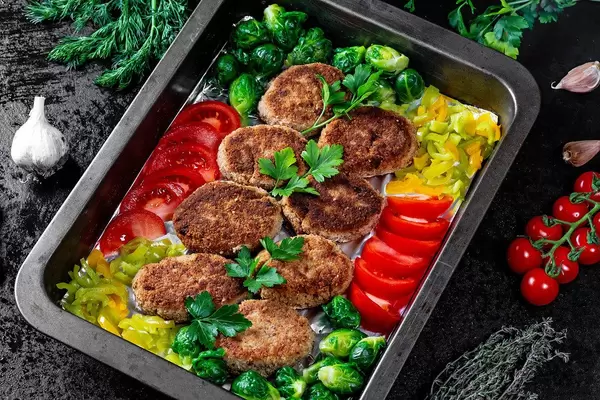 Saftige Frikadellen mit Rosenkohl, Tomatenscheiben, Paprika und Kräutern, auf einem dunklem Backblech im rustikalen Design