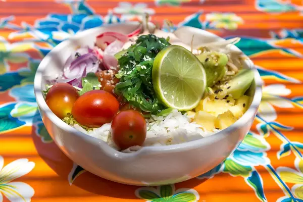 Salmon poke bowl with rice, lime, cherry tomatoes, radish, sprouts, avocado, herbs on a floral tablecloth