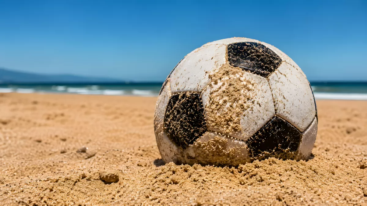 Sandbedeckter Fußball am Strand vor blauem Meer