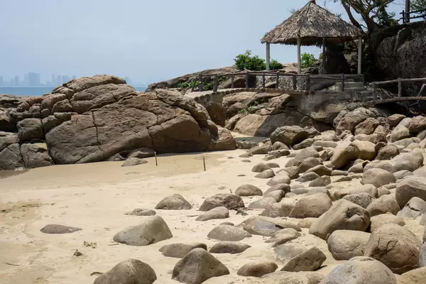 Sandstrand mit großen Steinen, Holzbrücke und kleiner Hütte mit Strohdach auf der Son Tra Halbinsel mit Blick auf die Skyline von Da Nang in Vietnam