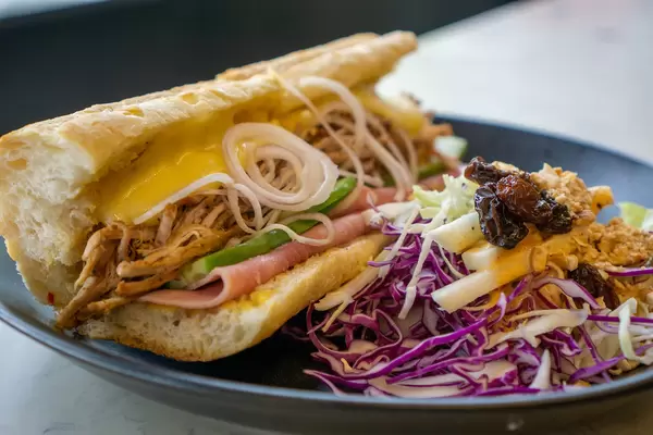 Sandwich mit Pulled Pork, Geschmolzenem Käse, Schinken, Zwiebeln und Gurken neben einem Krautsalat mit Rosinen und Erdnüssen Nahaufnahme