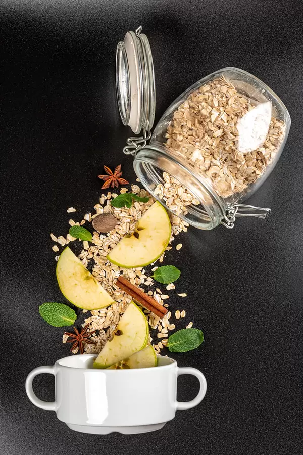 Scattered oatmeal from a jar with pieces of apple, spices and mint on a black background with a bowl, top view