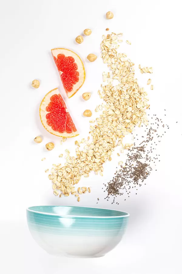 Scattered raw oatmeal, chia seeds, pomegranate pieces on a white plate with a bowl, top view