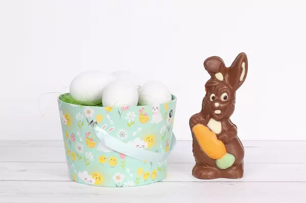 Schokoladen-Osterhase neben einem Eimer mit grünem Gras und weißen Ostereiern auf einem Holztisch mit weißem Hintergrund