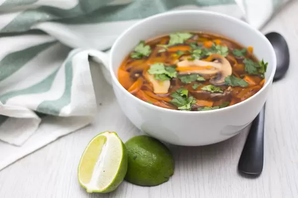 Schüssel mit asiatischer Suppe mit Pilzen, Karotten und Limette neben Löffel und Serviette