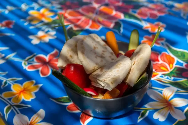 Schüssel mit Hummus, geschnittenem Gemüse und Pita auf Tischdecke mit Blumen-Muster