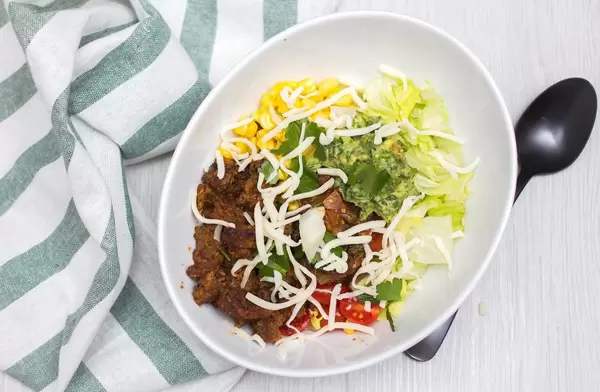 Schüssel Tacos mit Fleisch, Maiskörnern, Avocado-Dip und Salat neben Löffel und Serviette
