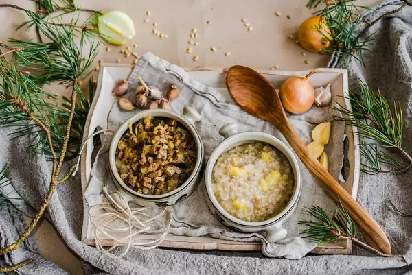 Schüsseln mit Perlgerstenbrei, Fleisch und Gemüse auf Holztablett mit Knoblauch und Zwiebeln