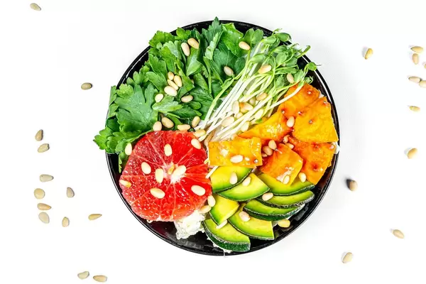 Schwarze Schale mit frischem Salat, Petersilie, Grapefruit, Mikrogrün, Avocado, Kiwano Horngurke und Pinienkernen von oben auf weißem Hintergrund fotografiert