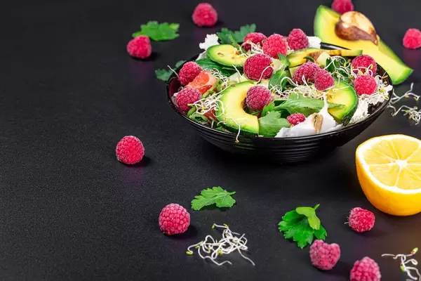 Schwarze Schale mit grünem Salat, Avocado-Scheiben, Himbeeren, Mikrogrün und anderen Zutaten auf schwarzem Hintergrund