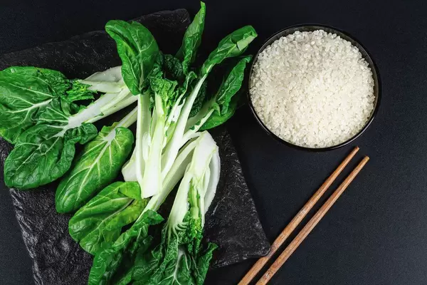 Schwarze Schüssel mit rohem Reis, Pak Choi auf einem schwarzen Schneidebrett und hölzernen Essstäbchen auf schwarzem Hintergrund