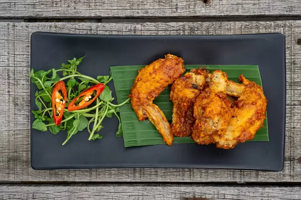 Schwarzer Teller auf einem Holztisch mit marinierten Chicken Wings auf einem Bananenblatt mit Chili und Kräuter von oben fotografiert