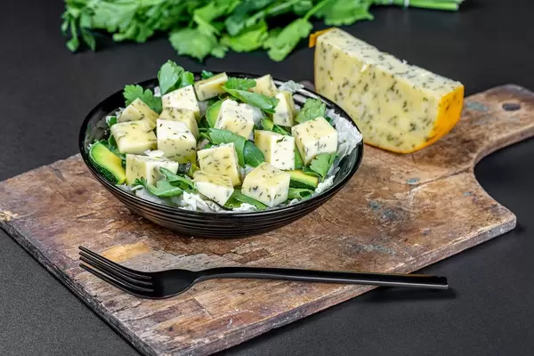 Schwarzer Teller mit frischem Salat, würzigen Käsewürfeln, Avocado-Scheiben und Petersilie auf einem Holzschneidebrett mit schwarzer Gabel und Stück Käse