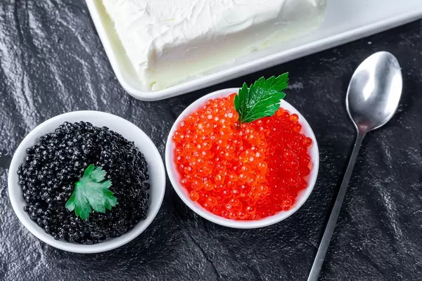 Schwarzer und roter Kaviar in kleinen Schüsseln, neben Butter und einem kleinen Löffel, auf einer schwarzen Steinplatte