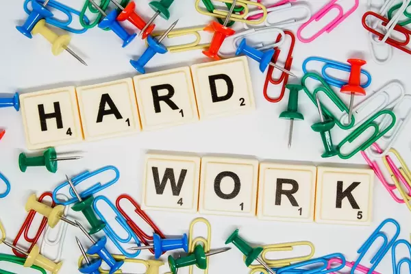 Scrabble-Steine legen das Wort "Hard Work" - Harte Arbeit, zwischen bunten Reißzwecken und Büroklammern