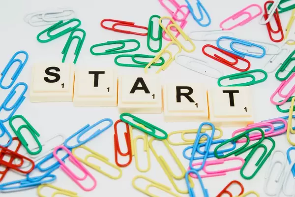 Scrabblesteine bilden das Wort Start, umgeben von bunten Büroklammern