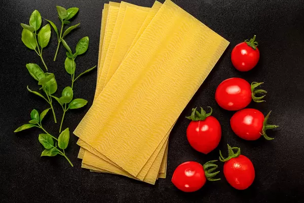 Sheets for lasagna with basil leaves and tomato on black, top view