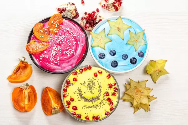 Sicht auf drei Schalem mit Haferflocken, frischen Sternfrüchten, Kaki, Beeren und Samen, auf einem weißen Holztisch