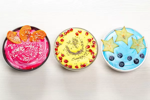 Sicht von oben auf bunte Frühstücks-Gerichte: Farbenfroher Joghurt in blau, rosa und gelb, mit Beeren und Früchten in Müslischüsseln