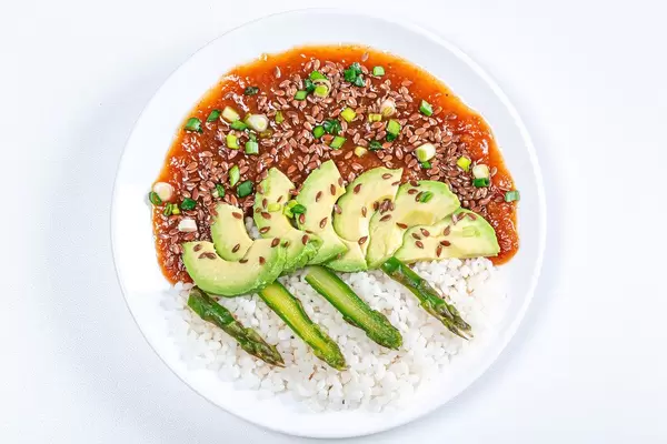 Sicht von oben auf einen Teller Reis mit Tomaten-Apfelsauce, Leinsamen, grüner Spargel und Avocado, vor weißem Hintergrund