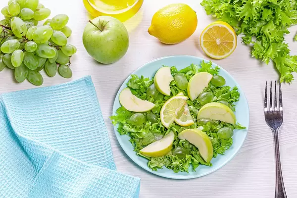 Sicht von oben auf einen vegetarischen Salat mit Äpfeln, Weintrauben und grünem Salat, auf einem weißen Tisch