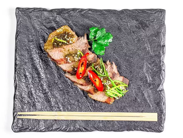 Sicht von oben auf geschnittene Filetstücke aus Rindfleisch, mit Chilischoten, Lauch und Sesamkernen, neben Essstäbchen auf einem schwarzen Schieferstein