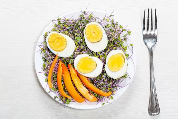 Sicht von oben auf hartgekochten Eiern, halbiert, auf Salat mit Microgreens und oranger Paprika, auf einem weißen Teller neben einer Gabel