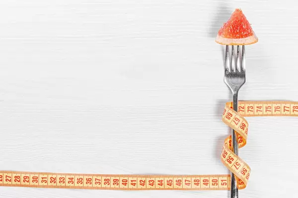 Slimming concept-a piece of grapefruit on a fork wrapped with measuring tape on a white wooden background