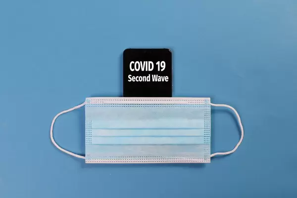 Smartphone with Covid-19 Second Wave text and medical face mask on blue bakcground