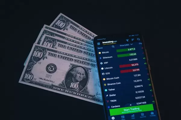 Smartphone zeigt Wert von Kryptowährungen neben amerikanischen Geldscheinen, vor schwarzem Hintergrund