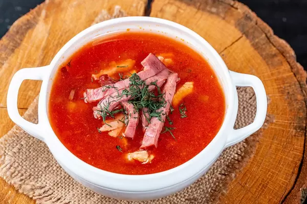 Soljanka Suppe mit geräuchertem Fleisch und Kräutern, in einer weißen Schale, im rustikalen Setting mit Baumstamm und Leinentuch