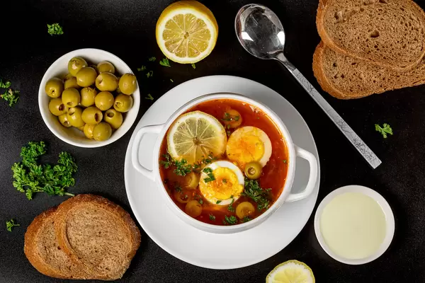 Solyanka soup on a dark background with olives, lemon, bread and herbs, top view