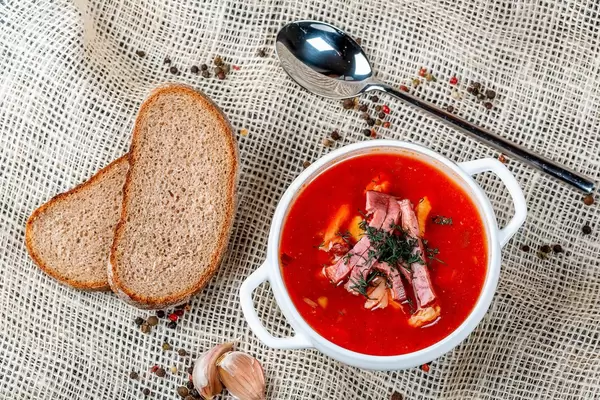 Soup saltwort with smoke meat, pickled cucumber, tomato sauce and greens on a background of burlap
