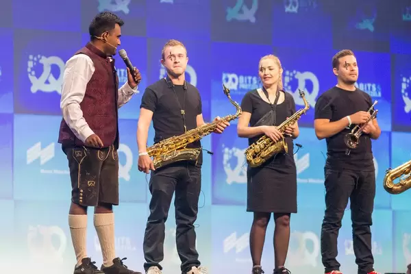 Speaker and Bits & Pretzels Host Dan Ram with Oktoberfest-look in lederhosen, next to trumpeters on stage
