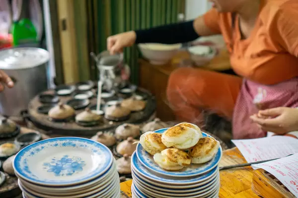 Steaming Hot Banh Can - Vietnamese Rice Cakes on a Plate with Woman preparing more Cakes in the Background