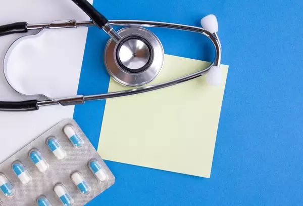 Stethoskop und Pillen auf blauem Hintergrund, mit leerem gelben Notizzettel