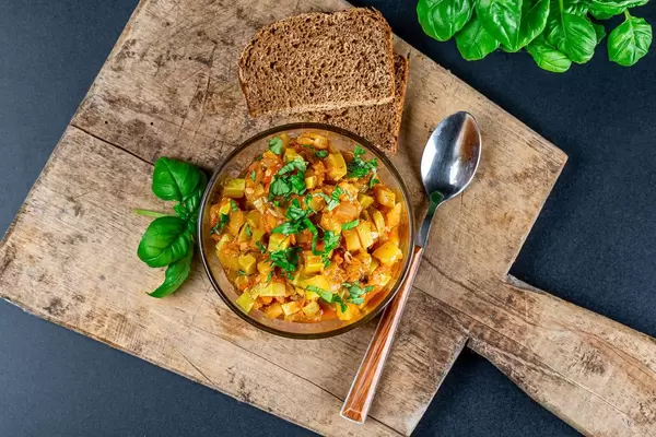 Stewed vegetables in a bowl on a dark background with fresh basil and slices of black bread, top view