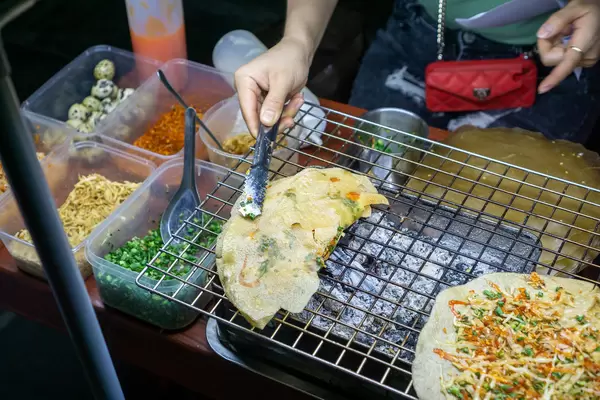 Street Food Seller preparing Vietnamese Pizza Banh Trang Nuong on a Barbecue Grill with Quail Eggs, Chili, Shrimps, Spring Onions, Fried Onions and Mayonnaise in Hoi An, Vietnam