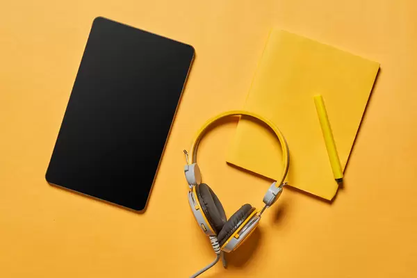 Tablet with a blank screen, yellow headphones and notepad on yellow background