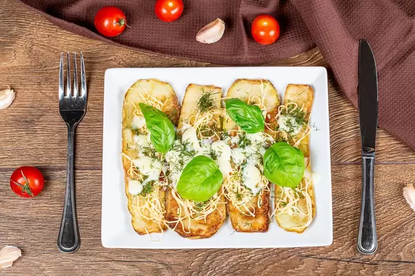 Tasty zucchini with cheese and herbs on a wooden background with knife and fork, top view