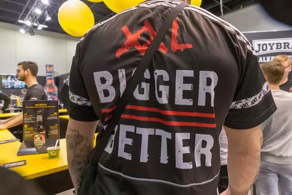 Tätowierter Mann im Bigger-Better T-Shirt steht vor dem Joybräu-Stand auf der Fibo-Messe Köln