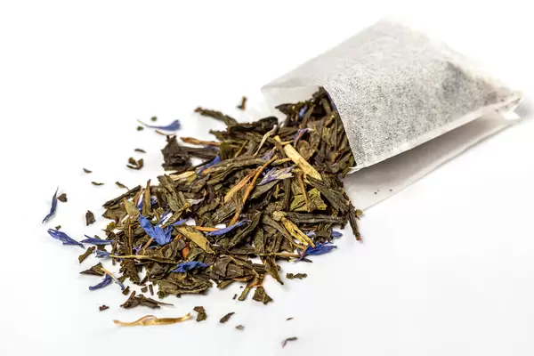 Tea bag with scattered green tea and blue flowers, close up