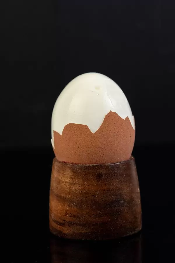 Teilweise geschältes Frühstücksei in Holzbecher vor spiegelndem schwarzem Hintergrund